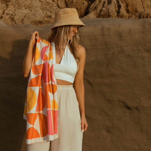 Woman enjoying a sunny day with a colorful Trek Light beach blanket and stylish hat against a rock backdrop.