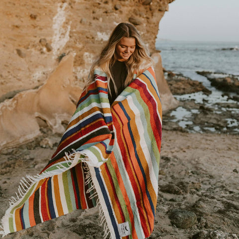 Woman wrapped in colorful striped Trek Light Halley's Comet Blanket by the rocky seaside, enjoying the view.