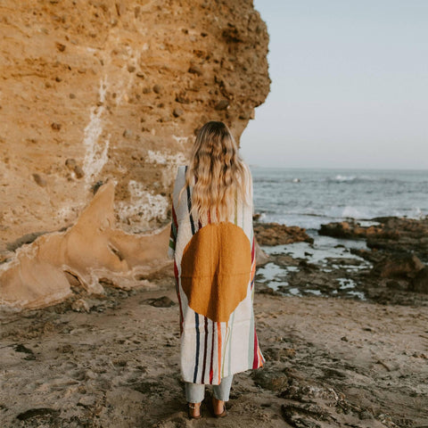 Person standing on rocky beach wrapped in a colorful blanket, gazing at the ocean.