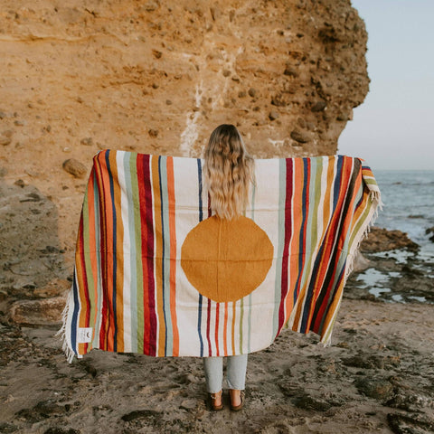 Woman holding a colorful striped blanket with a circular design, standing on a rocky beach. Trek Light inspired, handwoven beauty.