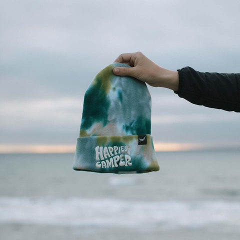 Hand holding Trek Light's Happiest Camper Tie Dye Beanie by the sea. Perfect for warmth, style, and spreading joy. 100% Cotton.