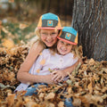 Two smiling children wearing Trek Light hats, sitting in a pile of autumn leaves near a tree. Perfect moment of outdoor joy.