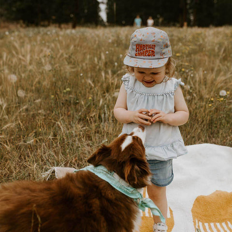 A smiling child wearing a 'Happiest Camper' kids hat plays in a meadow with a dog, embodying Trek Light adventure spirit.