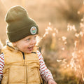 Smiling child wearing a Happiest Camper Kids Beanie by Trek Light, enjoying a sunny day outdoors.