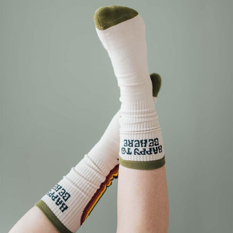 Person wearing "Happy To Be Here" socks in green and white with feet playfully raised, showcasing Trek Light comfort and style.