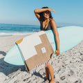 Smiling woman holding surfboard with Horizon Blanket on beach, highlighting sustainable Trek Light lifestyle.