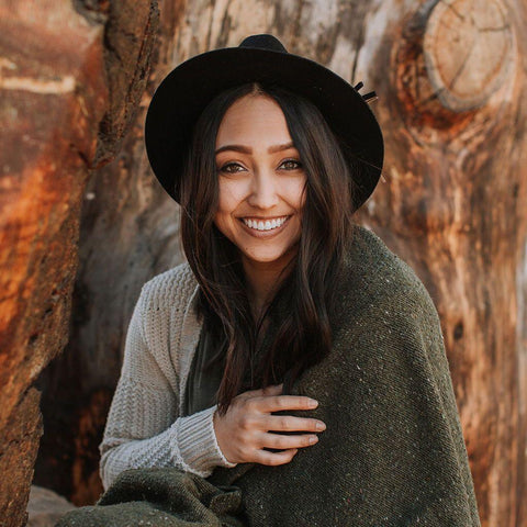 Woman smiling wrapped in Trek Light Humboldt Blanket near rustic tree