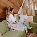 Cozy woman on green couch with Huntington Blanket, sand-colored and perfect for any adventure. Ideal for Trek Light outdoor fans.