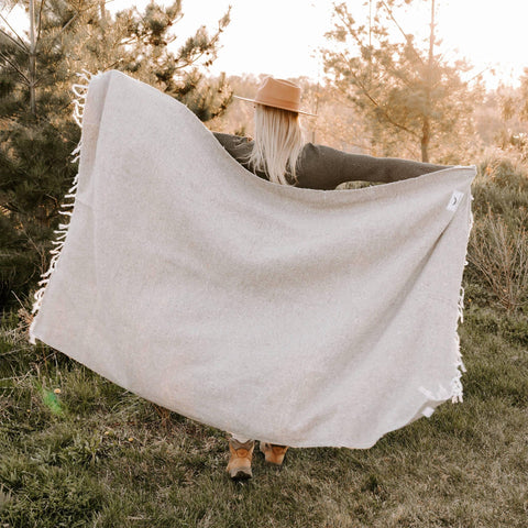 Woman holding a sand-colored Huntington Blanket in nature, perfect for beach days and cozy nights. Trek Light adventure vibes.