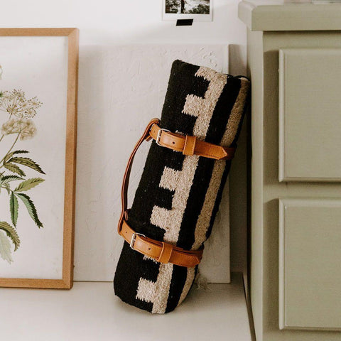 Trek Light handwoven blanket with leather carrier bundle, made from recycled and reclaimed materials, leaning stylishly on a dresser.