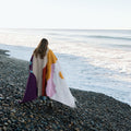 Cozy handwoven Mountain Sunrise Blanket made from recycled materials, draped over a person walking on a pebbled beach. By Trek Light.