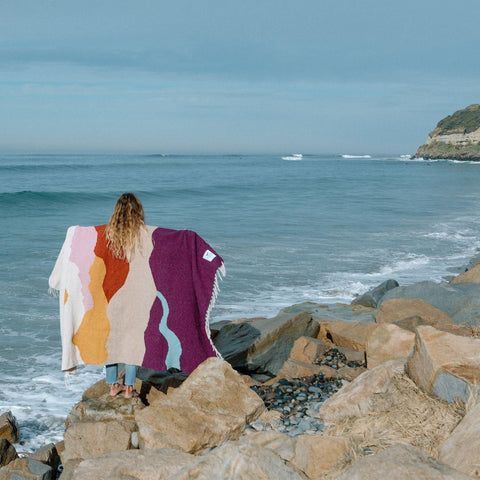 Person holding a handwoven Mountain Sunrise Blanket made from recycled clothing materials, standing on rocky shore. By Trek Light.