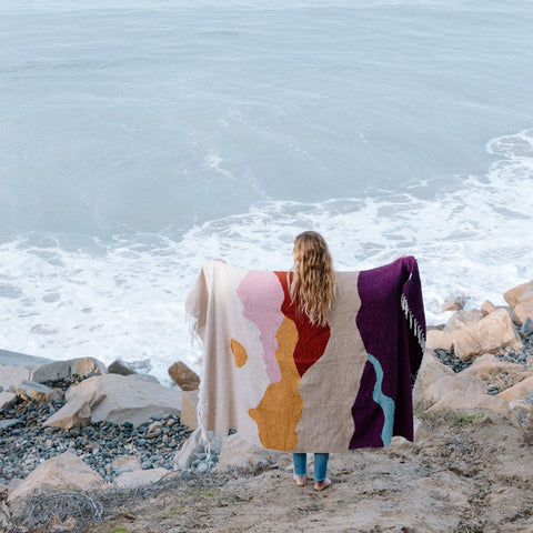 Person holding a cozy Mountain Sunrise Blanket, handwoven from recycled clothing, by the ocean. Beautiful and versatile. By Trek Light.