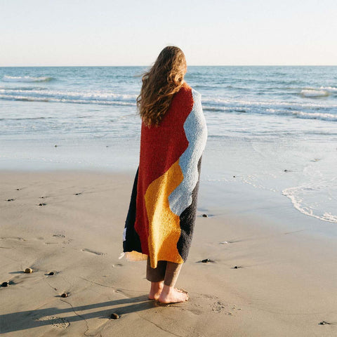 Woman wrapped in colorful Trek Light Mountains Blanket standing on sandy beach facing ocean waves.