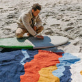 Man on beach admiring colorful Trek Light Mountains Blanket near surfboard, handwoven from recycled materials. Cozy and eco-friendly.