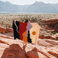 Person holding a colorful handwoven Mountains Blanket by Trek Light, set against a stunning mountain backdrop, made from recycled materials.