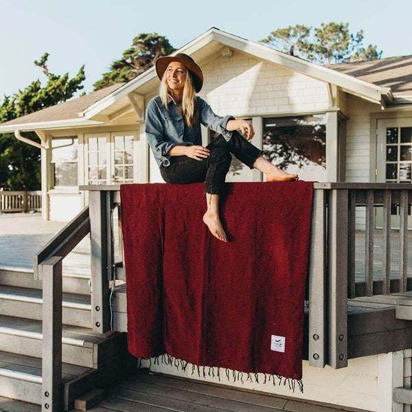 Woman in a hat sitting on a porch railing above a red Trek Light Napa Blanket, made from recycled materials. Eco-friendly cozy vibes.