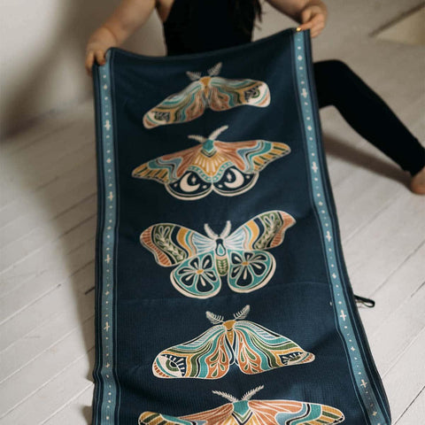 Colorful moth design on Trek Light tapestry held up by a person on a wooden floor.