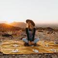 Woman sitting on a handwoven Peace Blanket from Trek Light at sunset, showcasing recycled clothing materials, versatile and cozy design.