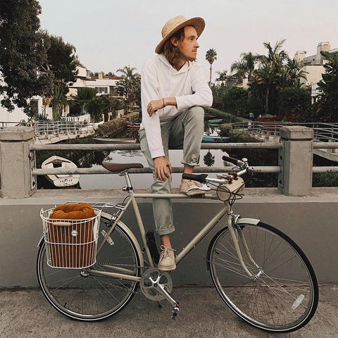 Person in a hat sitting on a vintage bike with a basket, with a Trek Light blanket conveniently stowed on their bike. 