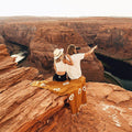 Couple sitting on cliff with a Trek Light Peace Blanket overlooking a canyon, enjoying a scenic view and pointing at the horizon.