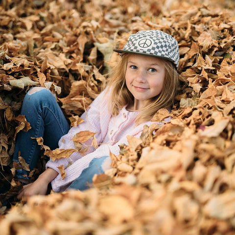 Child wearing a Peace Checkered Kids Hat by Trek Light, smiling and sitting in autumn leaves. Perfect accessory for cool kids.