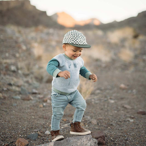 Toddler wearing a Peace Checkered Kids Hat with a Trek Light vibe, standing outdoors, feeling adventurous and cool in stylish denim and boots.