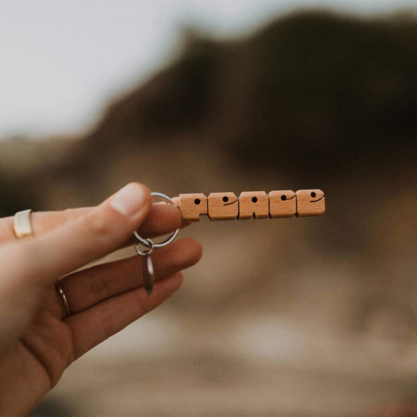 Hand holding Trek Light wooden PEACE keychain, perfect small gift with nickel split-ring hardware, rustic and charming design.