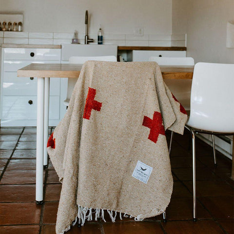 Trek Light Positive Vibrations Blanket draped over chair, handwoven with recycled materials, featuring red crosses and sustainable tag.