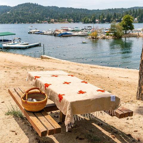 Trek Light Positive Vibrations Blanket on picnic bench by lake, showcasing sustainable, handwoven design with basket.