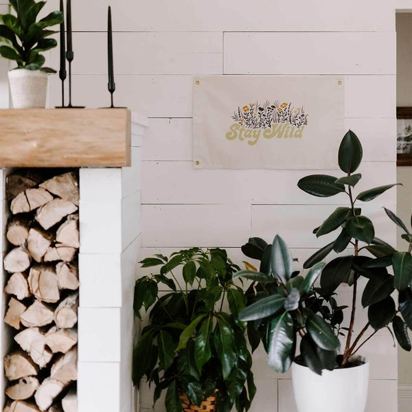 Stay Wild Canvas Flag by Trek Light hanging on a white wall with indoor plants, featuring hand-screened art and brass grommets.