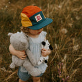 Child wearing colorful Trek Light Stay Wild hat, holding stuffed animals in a grassy field, ready for adventure.