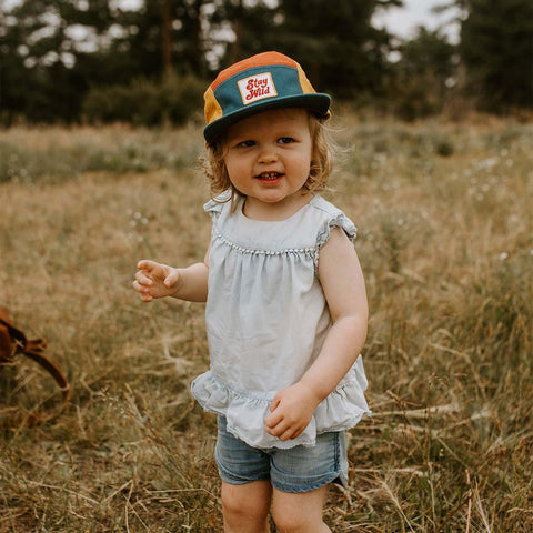 Child wearing colorful Trek Light Stay Wild hat, smiling in a field, ready for outdoor adventure. Perfect 5-panel camper style for kids.