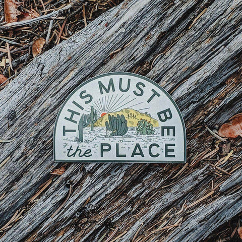 "This Must Be The Place" desert sticker by Trek Light on textured wood, showcasing cactus artwork and sunset vibes.