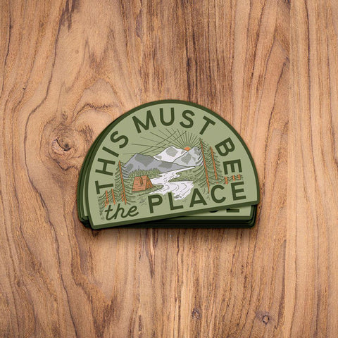 Trek Light 'This Must Be The Place' sticker featuring A-frame cabin, mountains, and river, on wooden background.