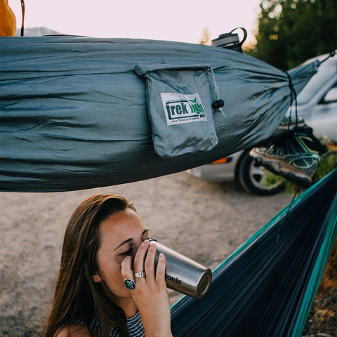 Woman enjoys a drink under a Trek Light VersaTrek hammock setup, showcasing its versatility as a gear loft, swing, and chair.