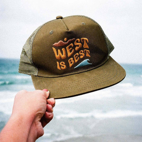 Trek Light's 'West Is Best' 5-panel trucker hat held by ocean view, ideal for adventures and rhyming swagger. Snapback, flat brim.