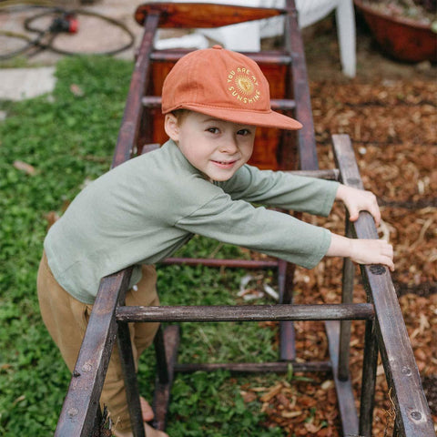 Child wearing Trek Light 'You Are My Sunshine' hat, playing outdoors with a rustic wooden frame, embodying adventure and joy.